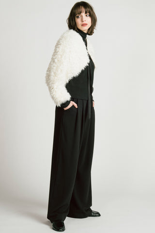Side view of woman in white studio, wearing black mock neck top, black wide leg pants and a white fuzzy bolero. 