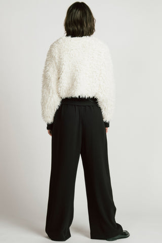 Back view of woman in white studio, wearing white fuzzy long sleeve top with black wide leg pants.