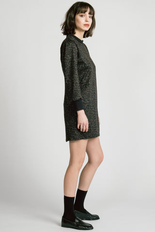 A side view of model wearing a black long-sleeve crewneck dress with leopard print, paired with black socks and black leather loafers, against a light gray studio background.