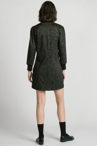 A back view of model wearing a black long-sleeve crewneck dress with leopard print, paired with black socks and black leather loafers, against a light gray studio background.