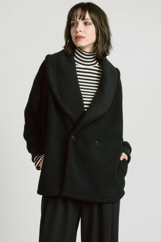 A woman wearing a black double-breasted jacket over a black and white striped turtleneck sweater and black trousers, photographed against a light gray studio background.