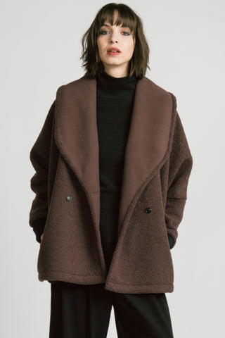 A woman wearing a brown double-breasted jacket over a black turtleneck sweater and black trousers, photographed against a light gray studio background.