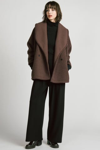 A woman wearing a brown double-breasted jacket over a black turtleneck sweater and black trousers, photographed against a light gray studio background.