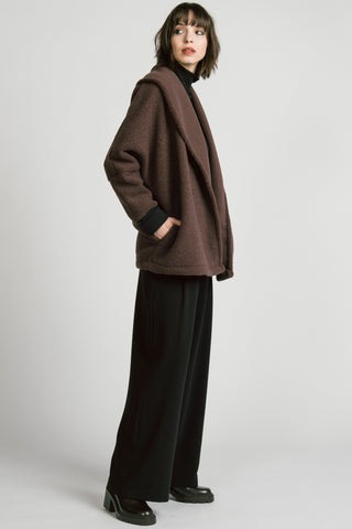 Side view of woman wearing a brown double-breasted jacket over a black turtleneck sweater and black trousers, photographed against a light gray studio background.