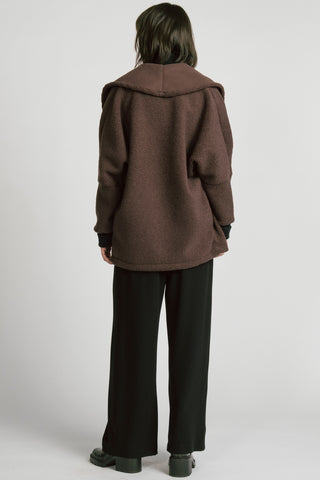 A woman shown from the back wearing a brown double-breasted jacket over a black turtleneck sweater and black trousers, photographed against a light gray studio background.