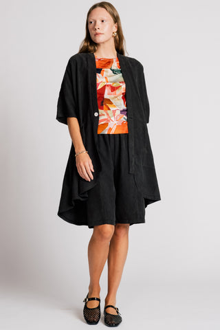 Woman in front of white background wearing wearing a black jacket over an orange printed blouse and black knee length shorts. 