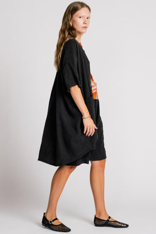 Woman in front of white background wearing wearing a black jacket over an orange printed blouse and black knee length shorts. 