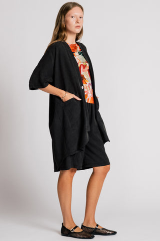 Woman in front of white background wearing wearing a black jacket over an orange printed blouse and black knee length shorts. 