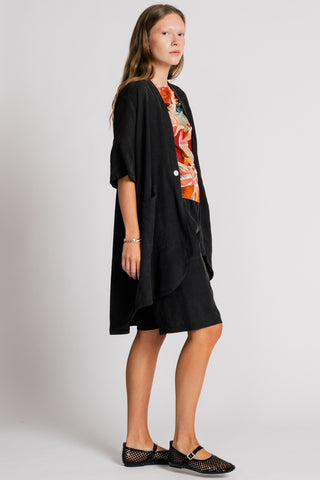 Woman in front of white background wearing wearing a black jacket over an orange printed blouse and black knee length shorts. 