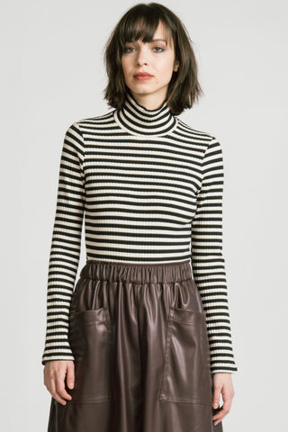 A woman wearing a black and off-white horizontally striped ribbed turtleneck sweater and a brown faux-leather skirt, standing against a light gray studio background.