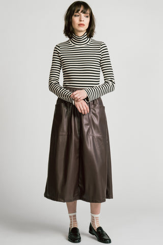 A model stands against a light gray studio background wearing a black and white horizontally striped mock-neck long-sleeve top, a brown faux-leather midi skirt with front patch pockets, patterned socks, and black leather loafers.