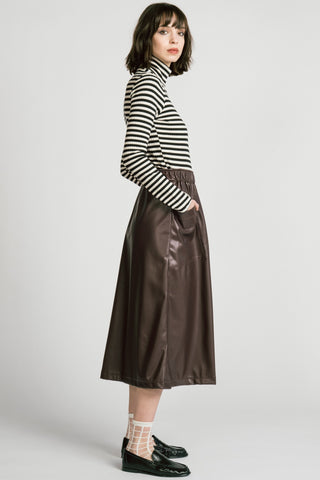 A model is shown from the side wearing a black and white horizontally striped long-sleeve mock neck top and a brown faux-leather midi skirt, paired with white socks and black loafers, against a light gray studio background.