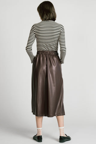 A model viewed from the back wearing a black and white horizontally striped long-sleeve top and a brown faux-leather midi skirt, paired with white socks and black shoes, against a light gray studio background.