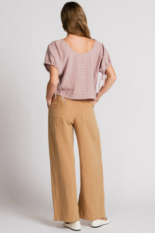 Back view of woman in front of white background wearing a pink gingham blouse and wide leg tan pants. 