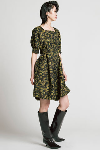 Woman in front of grey background wearing a gold and black floral jacquard babydoll dress with knee high black patent boots. 