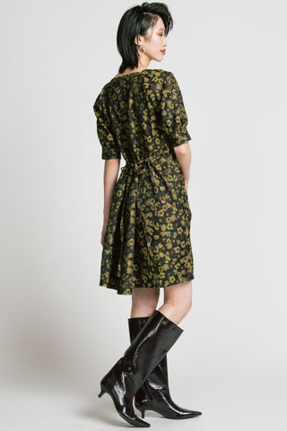 Back view of woman in front of grey background wearing a gold and black floral jacquard babydoll dress with knee high black patent boots. 
