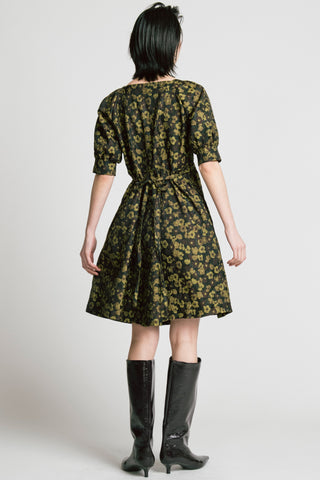Back view of woman in front of grey background wearing a gold and black floral jacquard babydoll dress with knee high black patent boots. 