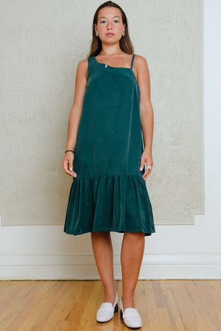 Woman in front of wall wearing green knee length dress with assymetrical neckline and gathered hem. 