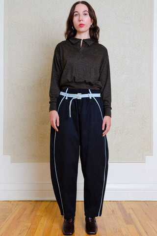 A woman stands indoors wearing a dark charcoal long-sleeve collared top, loose black barrel-leg pants with light blue piping and a matching belt, and dark brown shoes against a textured beige wall and wood floor.