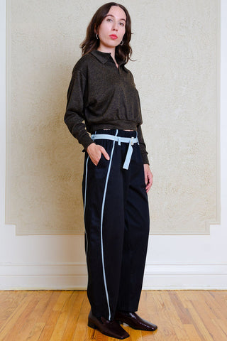 A woman stands indoors wearing a dark charcoal long-sleeve collared top, loose black barrel-leg pants with light blue piping and a matching belt, and dark brown shoes against a textured beige wall and wood floor.