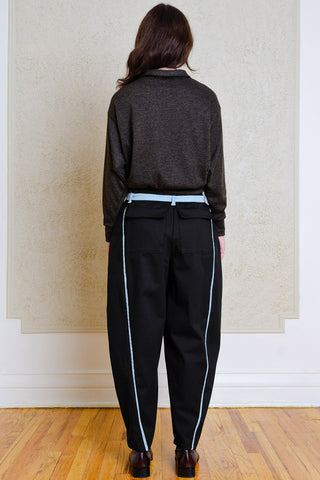 A person shown from the back wearing dark navy barrel-leg trousers with light blue side stripes, a dark charcoal knit sweater, and brown shoes, standing on a wooden floor against a beige wall.