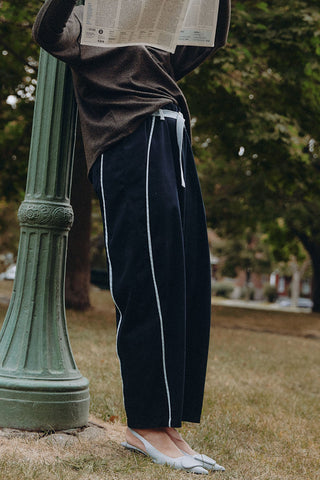 A person outdoors by a green lamppost wearing dark navy barrel-leg track pants with blue side stripes, a brown top, and white flats, holding a newspaper overhead.