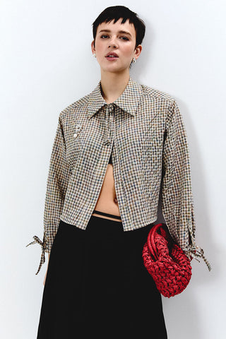 A model with short dark hair is wearing a cropped, long-sleeve button-up shirt in a beige and black gingham pattern, styled with a black skirt and a red woven handbag against a plain white background.