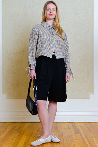 A woman stands in a room with a wood floor and textured beige wall, wearing a cropped multicolor gingham shirt, black shorts, and white sneakers, while holding a black shoulder bag.