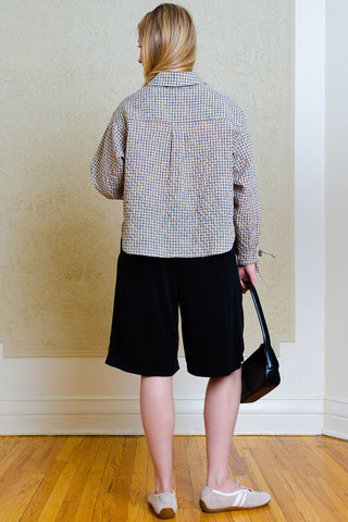 A woman shown from behind wearing a beige and brown checkered button-down shirt, black shorts, and white sneakers, holding a black handbag in an indoor setting with a textured beige wall and wooden floor.