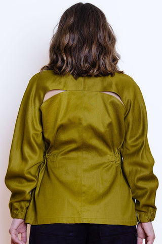 A woman viewed from the back wearing a khaki jacket with cutout details on the upper back and a elastic details at waist, paired with black pants.
