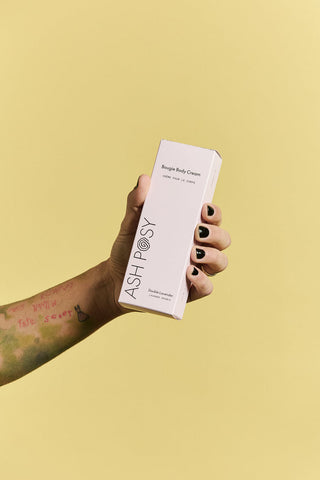 A hand with black nail polish and a tattoo holds a white rectangular box of ASH POSY Bougie Body Cream against a solid pale yellow background.