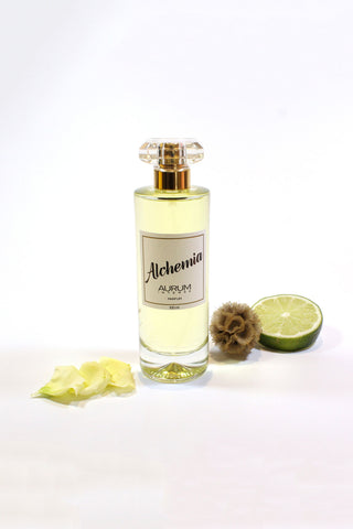 A clear glass 100ml Aurum Alchemia perfume bottle styled in front of a white background with white flower petals, a half a lime and dried flower. 