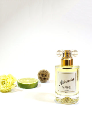 A clear glass 50ml Aurum Alchemia perfume bottle styled in front of a white background with white flower, a slice of lime and a dried flower. 