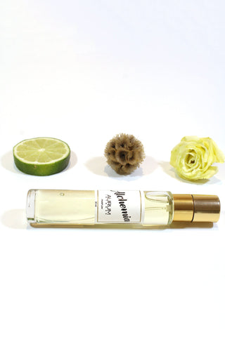 A clear glass 30ml Aurum Alchemia perfume bottle styled in front of a white background with white flower, dried flower and a slice of lime. 