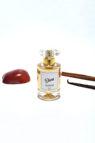 A 50ml bottle of Aurum Diva perfume in front of a white background with half a red apple, a stick of cinnamon and a vanilla bean. 