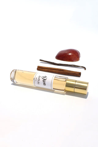 A 30ml bottle of Aurum Diva perfume in front of a white background with half a red apple, a stick of cinnamon and a vanilla bean. 