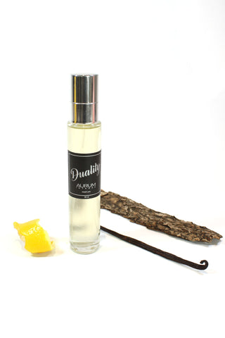 A 30ml square glass bottle of Aurum Duality perfume in front of white background, styled with lemon peel, vanilla pod and piece of wood. 