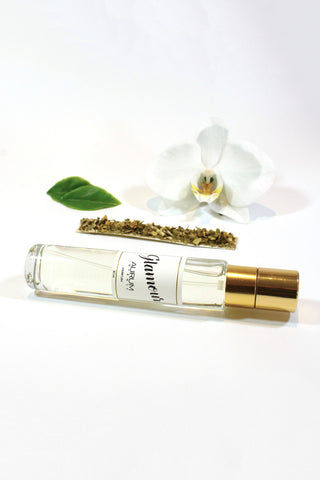 A clear Aurum Glamour perfume bottle lying on white background, with orchid flower, dried green tea and two green leaves. 