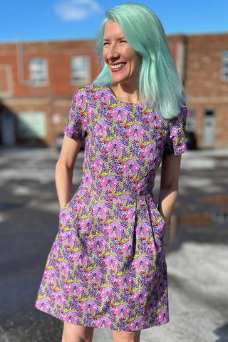Woman in front of brick building outside, wearing purple printed short sleeve structured dress. 