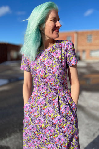 Woman in front of brick building outside, wearing purple printed short sleeve structured dress. 