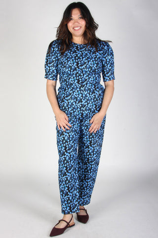Woman in front of grey background wearing blue mushroom print jumpsuit with 1/2 length sleeves. 