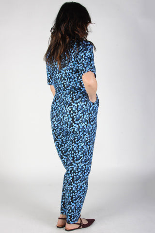 Back view of woman in front of grey background wearing blue mushroom print jumpsuit with 1/2 length sleeves. 