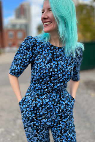 Woman with blue hair, standing outside, wearing blue print short sleeve jumpsuit. 