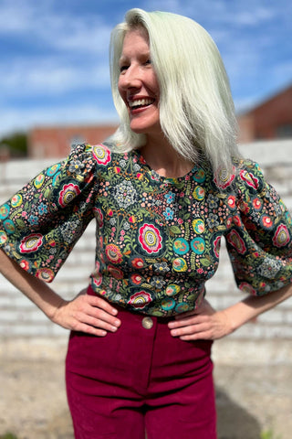 Woman standing outside, smiling, wearing red pants and floral print Paehen blouse by Birds of North America. 