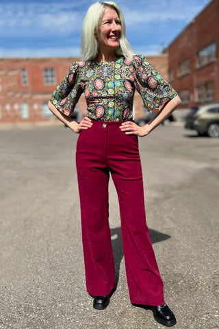 Woman standing outside, smiling, wearing red pants and floral print Paehen blouse by Birds of North America. 