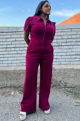 Woman outside in front of brick wall wearing mulberry zip up Tyrannulet jumpsuit by Birds of North America. 