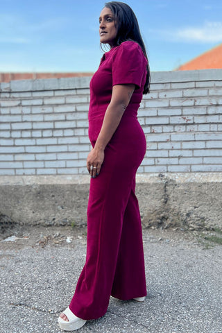 Side view of woman outside in front of brick wall wearing mulberry zip up Tyrannulet jumpsuit by Birds of North America. 