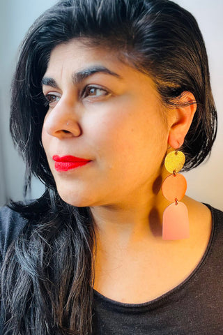 Woman wearing dangling leather earrings with gold and orange circle and pink arch. 