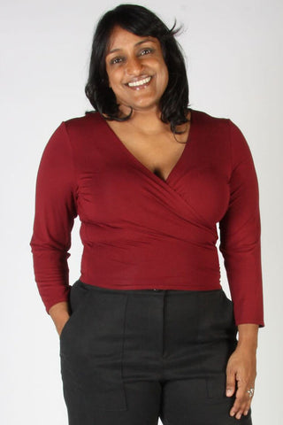 A smiling woman wearing a dark red long-sleeve wrap-style top and black trousers, standing against a plain light gray background.