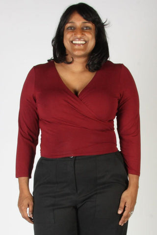 A woman smiling while wearing a long-sleeve wrap-style top in deep burgundy red and black trousers, standing against a plain light gray background.